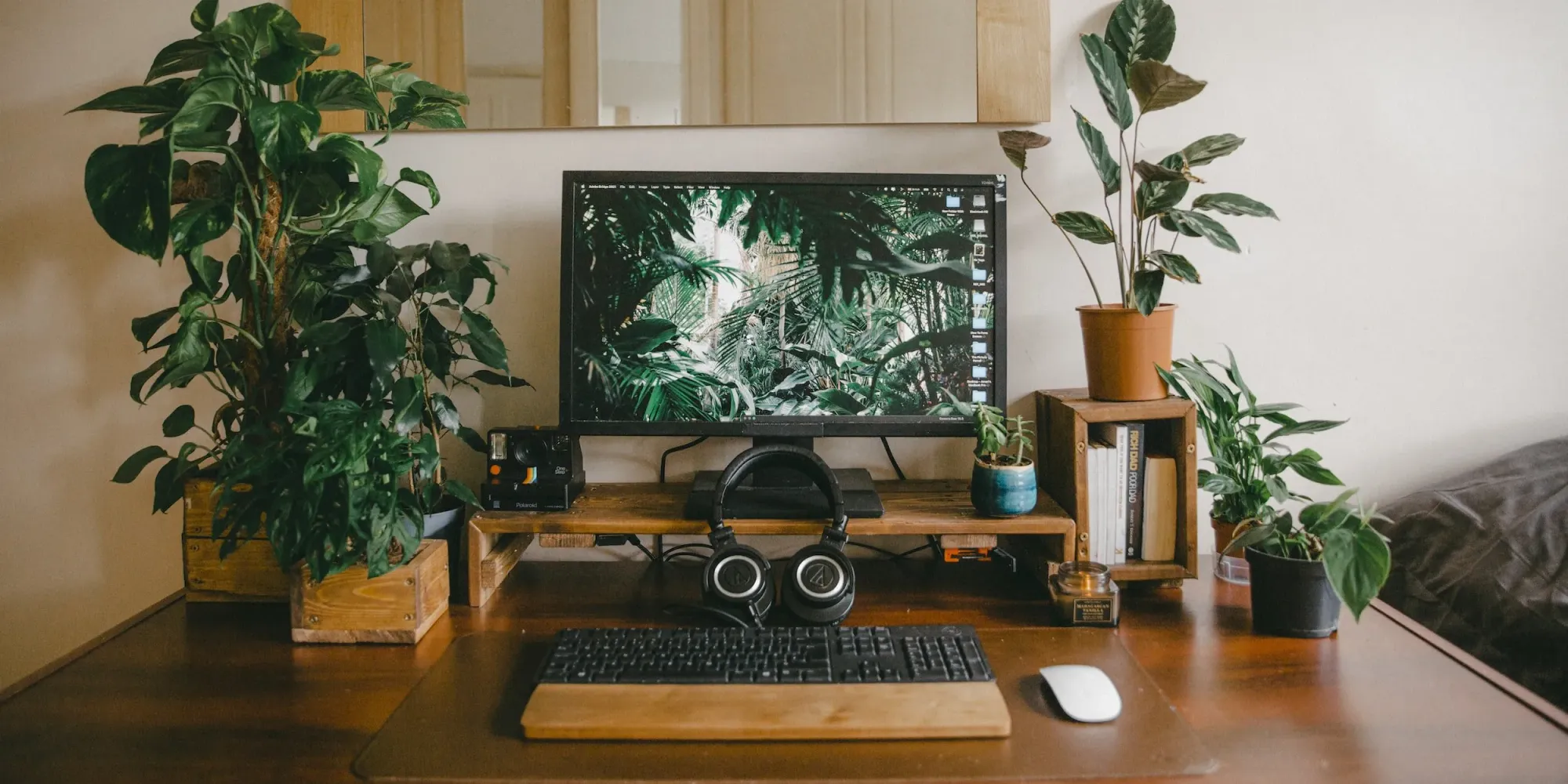 Plants, computer monitor, keyboard, headphones and mouse sitting on a wood desk with some books.